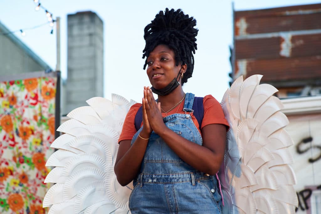 An actor wearing an orange tshirt and overalls with their hands in a prayer position. They are wearing a set of angel wings. 