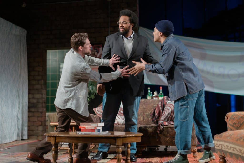 A photo of three actors in contemporaryy dress in a living room. The two actors on the side speak directly to the actor in the middle
