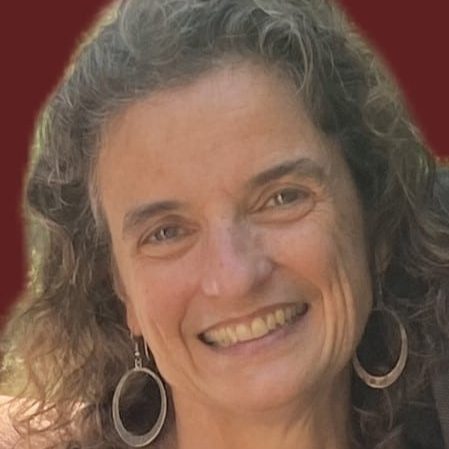 A photo of a woman in front of a red background. She has light brown hair and is wearing hoop earrings. 