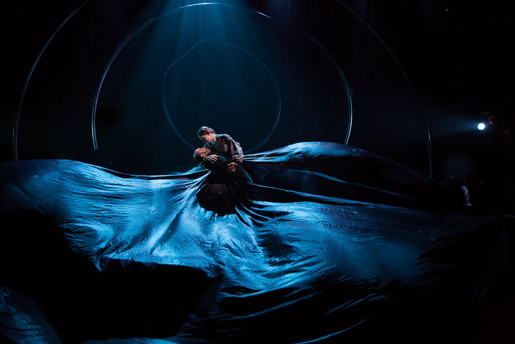 A production photo of a couple embracing. The woman is wearing a blue dress as wide as the stage, that looks like the sea.