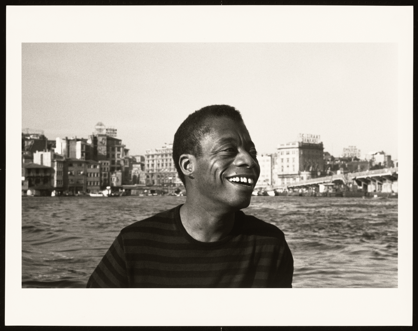 A photo of James Baldwin in a black tshirt with water behind him.